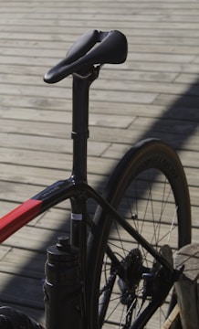 A close-up view of a bicycle focusing on the seat, seat post, and rear section of the frame and wheel. The bicycle has a sleek, modern design with a predominantly black frame and red accents. The background consists of wooden planks creating a rustic setting.