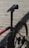 A close-up view of a bicycle focusing on the seat, seat post, and rear section of the frame and wheel. The bicycle has a sleek, modern design with a predominantly black frame and red accents. The background consists of wooden planks creating a rustic setting.