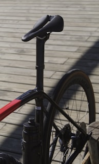 A close-up view of a bicycle focusing on the seat, seat post, and rear section of the frame and wheel. The bicycle has a sleek, modern design with a predominantly black frame and red accents. The background consists of wooden planks creating a rustic setting.