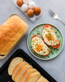 A top-down view of a breakfast arrangement with a loaf of bread, sliced bread on a tray, and a plate with two stuffed bell peppers topped with fried eggs and garnished with herbs. A carton containing both brown and white eggs is positioned nearby, along with a fork.