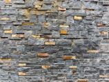 A textured stone wall composed of rectangular slate tiles with varying shades. The stones are stacked horizontally, showcasing a mix of natural grey and brown tones.