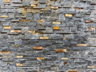 A textured stone wall composed of rectangular slate tiles with varying shades. The stones are stacked horizontally, showcasing a mix of natural grey and brown tones.