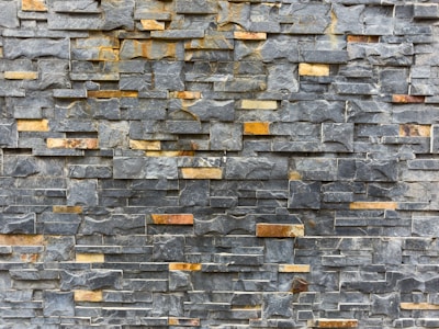 A textured stone wall composed of rectangular slate tiles with varying shades. The stones are stacked horizontally, showcasing a mix of natural grey and brown tones.