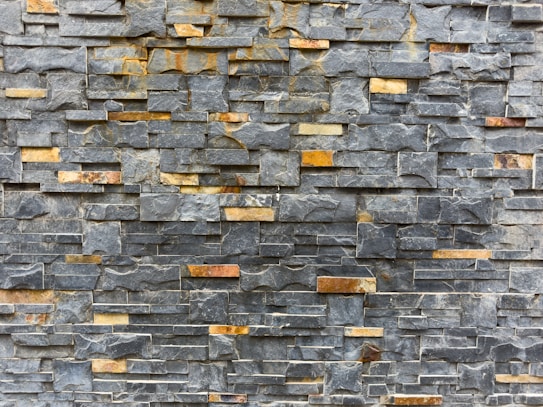 A textured stone wall composed of rectangular slate tiles with varying shades. The stones are stacked horizontally, showcasing a mix of natural grey and brown tones.