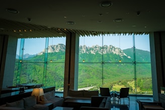 A spacious and modern interior with large glass windows providing a panoramic view of a mountainous landscape. The room is furnished with comfortable seating, tables, and soft lighting, while the striking mountain range outside is lush with greenery and rocky peaks.