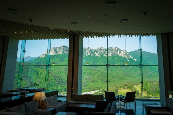 A spacious and modern interior with large glass windows providing a panoramic view of a mountainous landscape. The room is furnished with comfortable seating, tables, and soft lighting, while the striking mountain range outside is lush with greenery and rocky peaks.