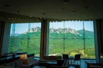 A spacious and modern interior with large glass windows providing a panoramic view of a mountainous landscape. The room is furnished with comfortable seating, tables, and soft lighting, while the striking mountain range outside is lush with greenery and rocky peaks.