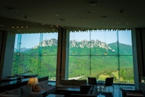 A spacious and modern interior with large glass windows providing a panoramic view of a mountainous landscape. The room is furnished with comfortable seating, tables, and soft lighting, while the striking mountain range outside is lush with greenery and rocky peaks.