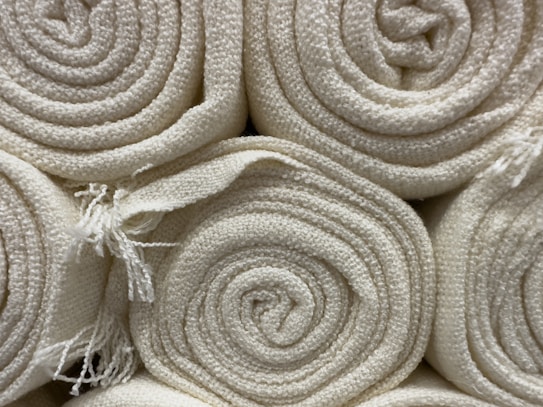 Neatly rolled stacks of off-white textured fabric or cloth are arranged closely together. Frayed edges are slightly visible, giving a natural and raw appearance.