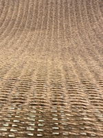A close-up view of a woven texture, likely made from natural fibers such as rattan or wicker. The pattern consists of interlaced strands creating a grid-like, uniform appearance with a slight curve.