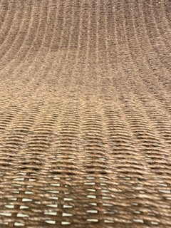 A close-up view of a woven texture, likely made from natural fibers such as rattan or wicker. The pattern consists of interlaced strands creating a grid-like, uniform appearance with a slight curve.