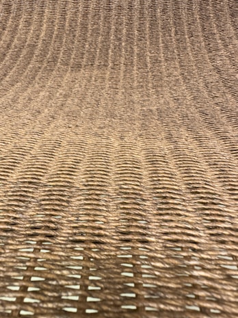 A close-up view of a woven texture, likely made from natural fibers such as rattan or wicker. The pattern consists of interlaced strands creating a grid-like, uniform appearance with a slight curve.