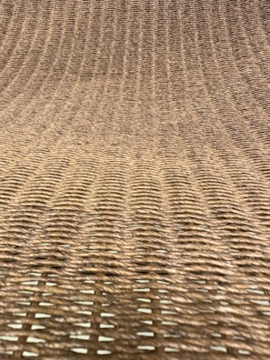 A close-up view of a woven texture, likely made from natural fibers such as rattan or wicker. The pattern consists of interlaced strands creating a grid-like, uniform appearance with a slight curve.