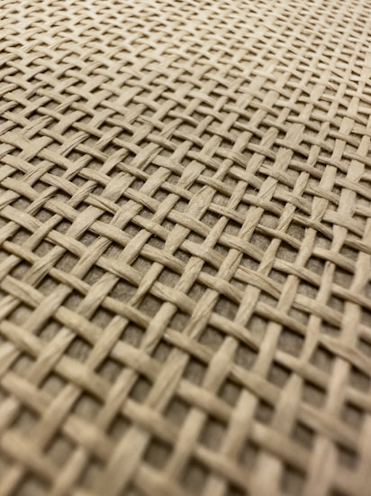 A close-up view of a woven material featuring an interlaced pattern of light brown fibers crisscrossing in a uniform grid formation. The texture appears natural, possibly made of wood or rattan, with a slightly rough surface.
