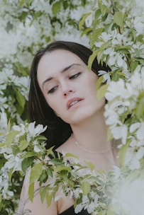 A woman is partially surrounded by lush green leaves and white blossoms, creating a serene and natural setting. Her expression appears contemplative, and she is wearing a simple necklace.