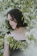 A woman with short, dark hair is surrounded by lush green leaves and white flowers. She is wearing a black sleeveless top and a delicate necklace. Her tattoo is visible on her arm, and she appears to be in a calm and serene state, gently touching the flowers.