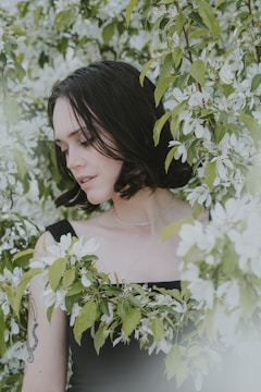 A woman with short, dark hair is surrounded by lush green leaves and white flowers. She is wearing a black sleeveless top and a delicate necklace. Her tattoo is visible on her arm, and she appears to be in a calm and serene state, gently touching the flowers.