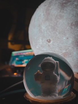 A small astronaut figurine is encased inside a glass sphere, with a blurred view of a moon-like object in the background. The sphere reflects dim lighting, adding an ethereal glow. The scene is accompanied by an out-of-focus computer screen displaying colorful light patterns.