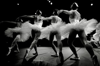 A group of ballet dancers in mid-performance, wearing elegant tutus and pointe shoes. The dancers are gracefully linked, displaying a sense of unity and fluidity in their movements. The lighting highlights the texture of the costumes and the muscular definition of the dancers, with a dramatic focus on their poised poses.
