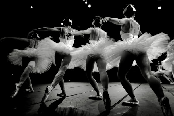 A group of ballet dancers in mid-performance, wearing elegant tutus and pointe shoes. The dancers are gracefully linked, displaying a sense of unity and fluidity in their movements. The lighting highlights the texture of the costumes and the muscular definition of the dancers, with a dramatic focus on their poised poses.