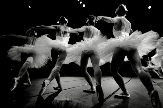 A group of ballet dancers in mid-performance, wearing elegant tutus and pointe shoes. The dancers are gracefully linked, displaying a sense of unity and fluidity in their movements. The lighting highlights the texture of the costumes and the muscular definition of the dancers, with a dramatic focus on their poised poses.