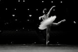 A ballet dancer gracefully poses on a stage, wearing a tutu and performing an arabesque. The background is dimly lit, with empty rows of theater seats visible.