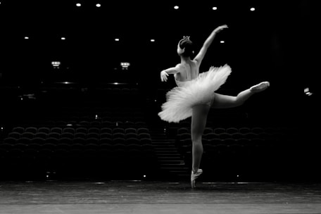 A ballet dancer gracefully poses on a stage, wearing a tutu and performing an arabesque. The background is dimly lit, with empty rows of theater seats visible.