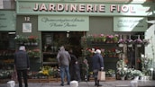 A storefront with a sign that reads 'Jardinerie Fiol' is visible, displaying a variety of plants and flowers. Several people are browsing the flower pots and hanging plants. The storefront has a green facade with decorative elements, and there is signage in French providing details about the store's offerings and services.