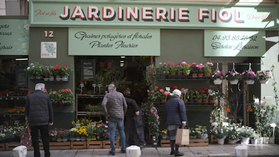 A storefront with a sign that reads 'Jardinerie Fiol' is visible, displaying a variety of plants and flowers. Several people are browsing the flower pots and hanging plants. The storefront has a green facade with decorative elements, and there is signage in French providing details about the store's offerings and services.