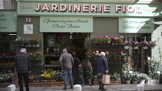 A storefront with a sign that reads 'Jardinerie Fiol' is visible, displaying a variety of plants and flowers. Several people are browsing the flower pots and hanging plants. The storefront has a green facade with decorative elements, and there is signage in French providing details about the store's offerings and services.