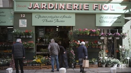 A storefront with a sign that reads 'Jardinerie Fiol' is visible, displaying a variety of plants and flowers. Several people are browsing the flower pots and hanging plants. The storefront has a green facade with decorative elements, and there is signage in French providing details about the store's offerings and services.