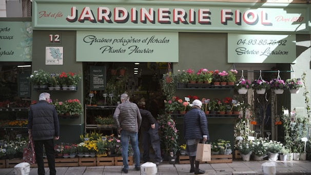 A storefront with a sign that reads 'Jardinerie Fiol' is visible, displaying a variety of plants and flowers. Several people are browsing the flower pots and hanging plants. The storefront has a green facade with decorative elements, and there is signage in French providing details about the store's offerings and services.