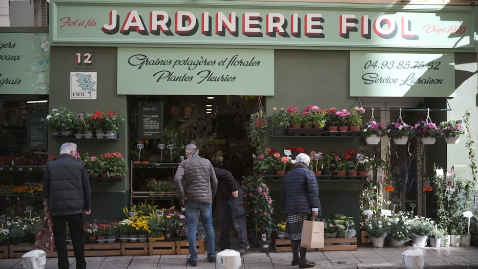 A storefront with a sign that reads 'Jardinerie Fiol' is visible, displaying a variety of plants and flowers. Several people are browsing the flower pots and hanging plants. The storefront has a green facade with decorative elements, and there is signage in French providing details about the store's offerings and services.