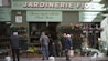 A storefront with a sign that reads 'Jardinerie Fiol' is visible, displaying a variety of plants and flowers. Several people are browsing the flower pots and hanging plants. The storefront has a green facade with decorative elements, and there is signage in French providing details about the store's offerings and services.