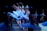 A group of ballerinas in white tutus gracefully perform on stage, extending their arms and legs in a synchronized dance formation under dim blue lighting.