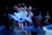 A group of ballerinas in white tutus gracefully perform on stage, extending their arms and legs in a synchronized dance formation under dim blue lighting.