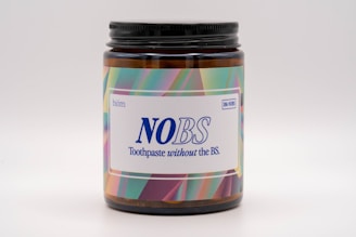 A brown jar with a black lid featuring a brightly colored label. The label displays the text 'NOBS' in large blue letters, along with the slogan 'Toothpaste without the BS.' The background of the label has a colorful, abstract pattern including shades of green, pink, and purple.