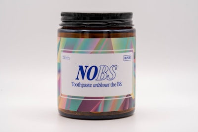 A brown jar with a black lid featuring a brightly colored label. The label displays the text 'NOBS' in large blue letters, along with the slogan 'Toothpaste without the BS.' The background of the label has a colorful, abstract pattern including shades of green, pink, and purple.