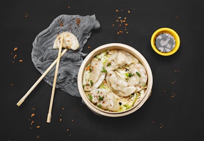 Several dumplings are arranged in a bamboo steamer filled with shredded cabbage and garnished with chopped green onions and seasoning. A pair of chopsticks holds one dumpling above a textured gray cloth, while scattered crumbs and a small yellow bowl of soy sauce add detail to the composition, set against a black background.