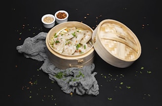 A bamboo steamer filled with dumplings garnished with chopped herbs is placed against a dark background. Next to the steamer, there are small white bowls containing sesame seeds and chili flakes. A grey textured cloth is also present around the steamer.