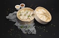 A bamboo steamer filled with dumplings garnished with chopped herbs is placed against a dark background. Next to the steamer, there are small white bowls containing sesame seeds and chili flakes. A grey textured cloth is also present around the steamer.