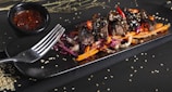 A rectangular black plate holds a colorful dish featuring tender pieces of grilled meat topped with sesame seeds and sliced red chili peppers. The meat is rested on a bed of vibrant shredded red cabbage, carrots, and onion. A black bowl of red sauce is placed nearby, and sesame seeds are scattered across the surface.