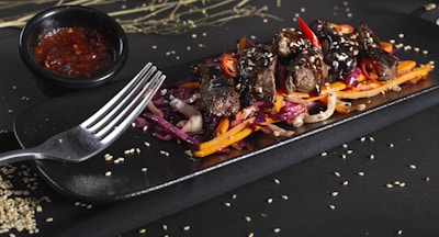A rectangular black plate holds a colorful dish featuring tender pieces of grilled meat topped with sesame seeds and sliced red chili peppers. The meat is rested on a bed of vibrant shredded red cabbage, carrots, and onion. A black bowl of red sauce is placed nearby, and sesame seeds are scattered across the surface.