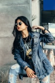 A fashionable individual wearing a denim jacket and sunglasses poses with confidence. The person has long dark hair and is adorned with jewelry, including earrings and a wristwatch. They appear to be sitting on a concrete step, with potted plants and cones visible in the background.