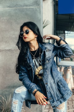 A fashionable individual wearing a denim jacket and sunglasses poses with confidence. The person has long dark hair and is adorned with jewelry, including earrings and a wristwatch. They appear to be sitting on a concrete step, with potted plants and cones visible in the background.