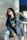 A fashionable individual wearing a denim jacket and sunglasses poses with confidence. The person has long dark hair and is adorned with jewelry, including earrings and a wristwatch. They appear to be sitting on a concrete step, with potted plants and cones visible in the background.