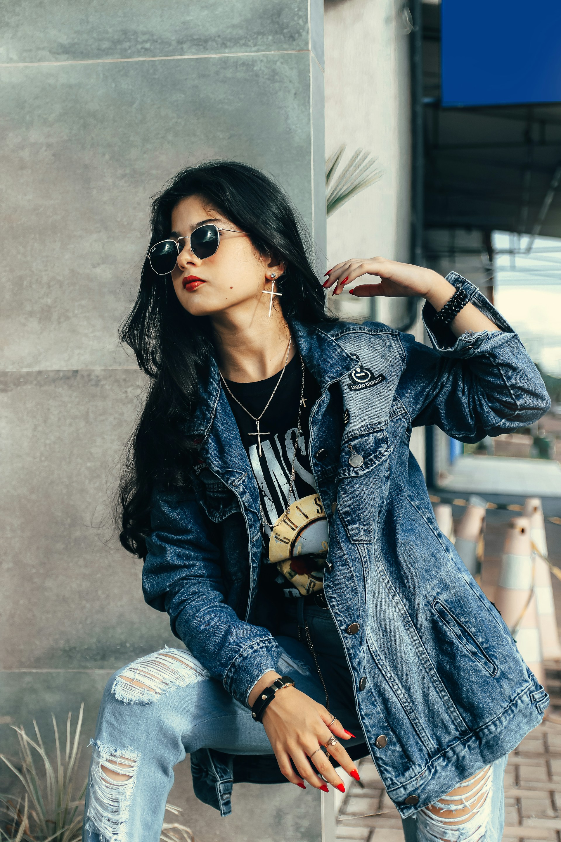 A fashionable individual wearing a denim jacket and sunglasses poses with confidence. The person has long dark hair and is adorned with jewelry, including earrings and a wristwatch. They appear to be sitting on a concrete step, with potted plants and cones visible in the background.