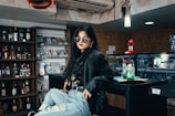 A person with long dark hair and sunglasses leans casually against a bar or counter. They wear a black leather jacket and distressed jeans, exuding a chic, laid-back style. Behind them is a wall lined with alcohol bottles and a display of magazines. The environment is warmly lit, suggesting a cozy and stylish setting.