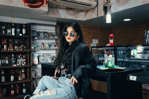 A person with long dark hair and sunglasses leans casually against a bar or counter. They wear a black leather jacket and distressed jeans, exuding a chic, laid-back style. Behind them is a wall lined with alcohol bottles and a display of magazines. The environment is warmly lit, suggesting a cozy and stylish setting.