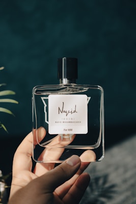 A hand holds a clear glass perfume bottle with a black cap. The bottle has a label with stylized text reading 'Navid Soir' and 'For Him' in smaller print, set against a dark, blurred background with hints of green foliage.